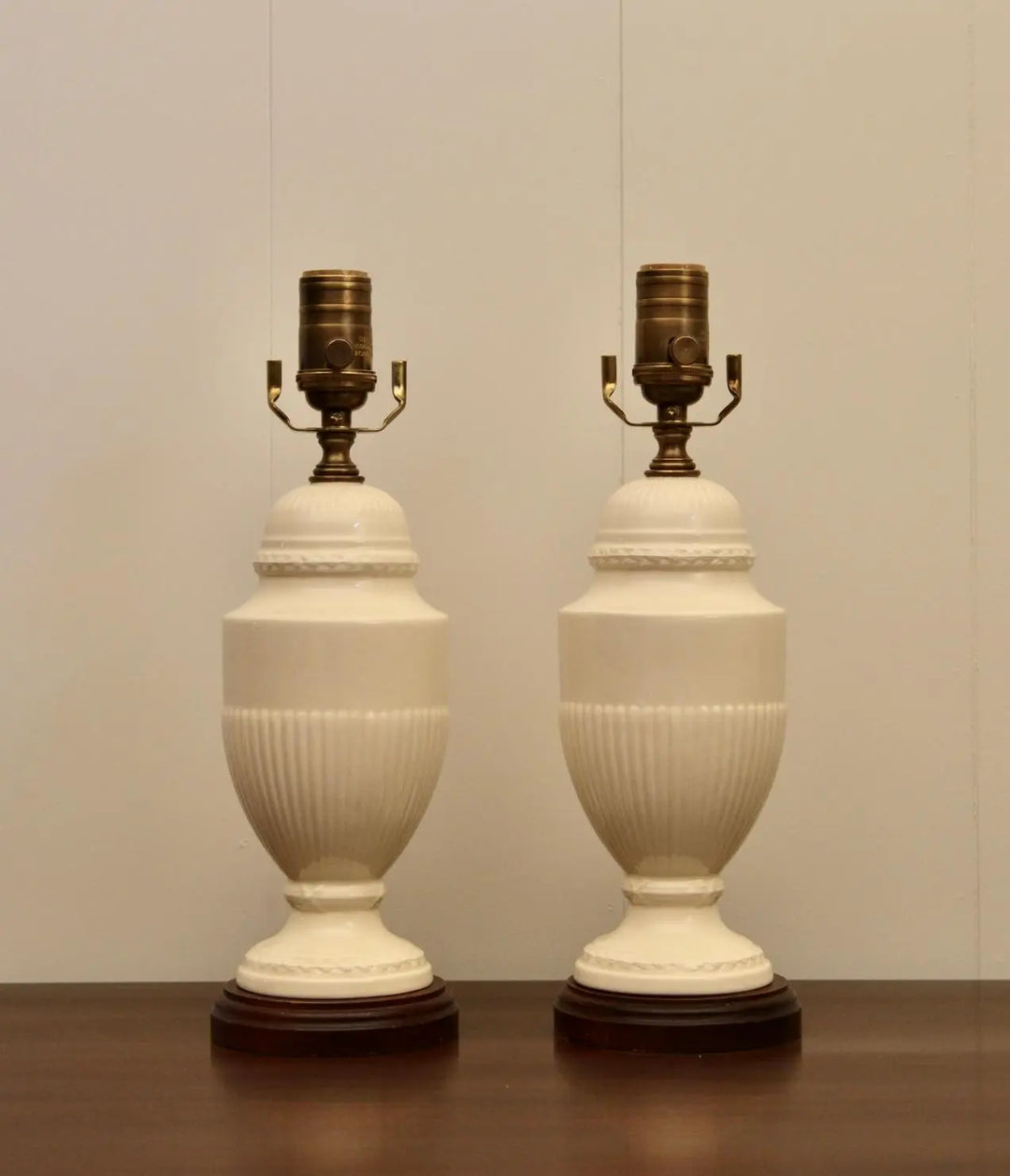 Pair of classic vintage cream ceramic table lamps with brass fittings and decorative bases on a wooden table.