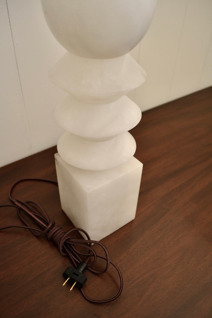 Close-up of white alabaster table lamp featuring stacked diamond shapes and a cube base, with cord wrapped beside it.
