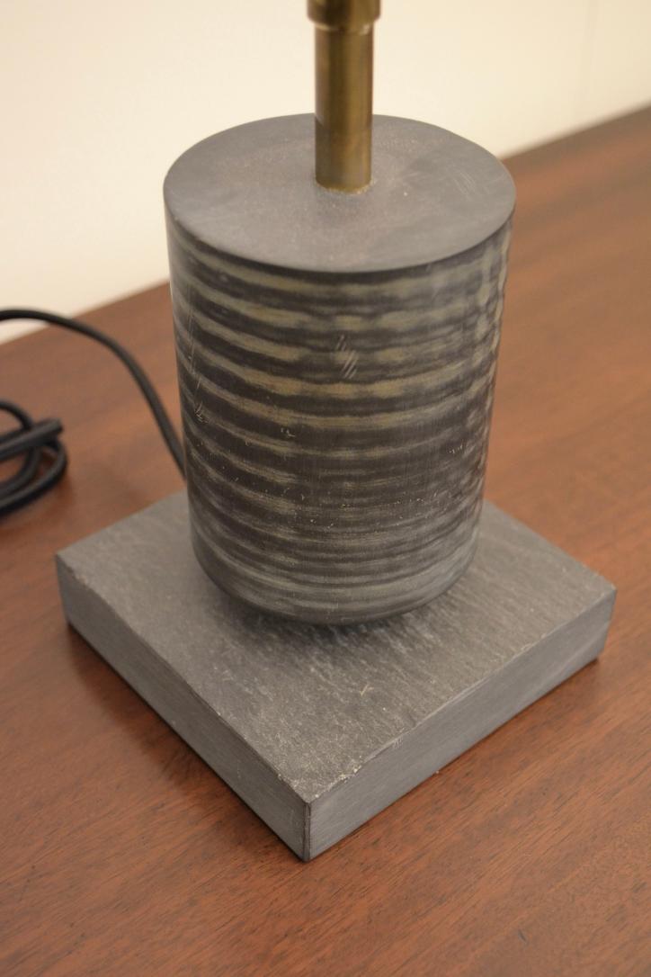 Close-up of a black ridged cylindrical lamp base on a square slate platform with a brass rod